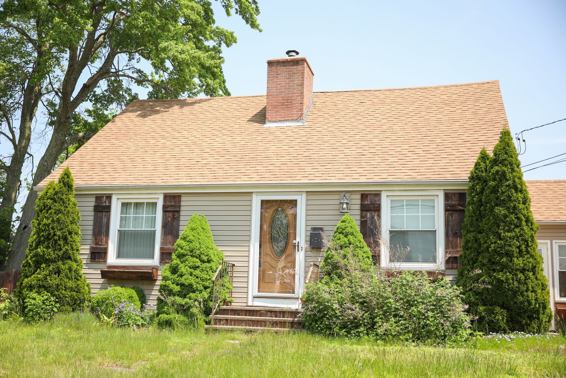 Small beige house with brown roof, shutters, chimney, and landscaping, blue sky in background.