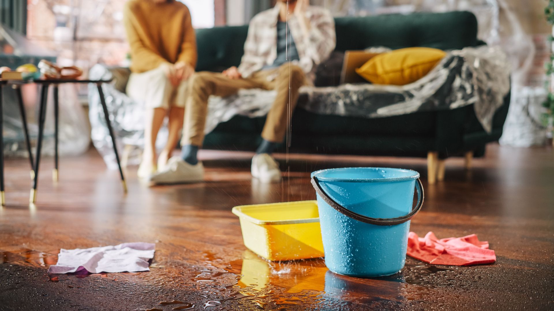 Two people on a sofa, looking distressed, as water floods a room with buckets on the floor.