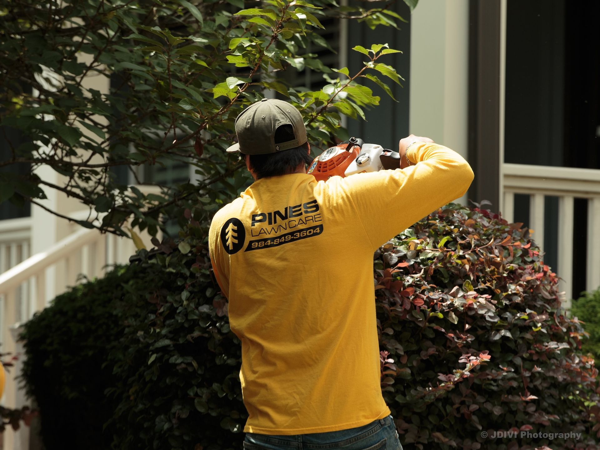 Man in yellow shirt trimming a bush with a hedge trimmer.