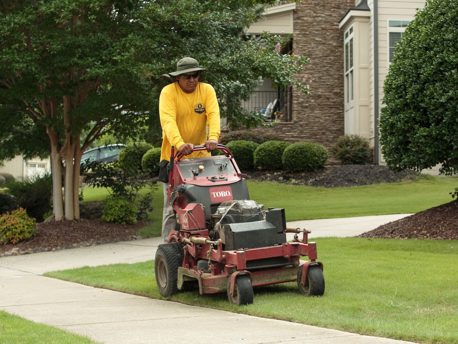 Man in yellow shirt mowing grass with red mower on sidewalk.