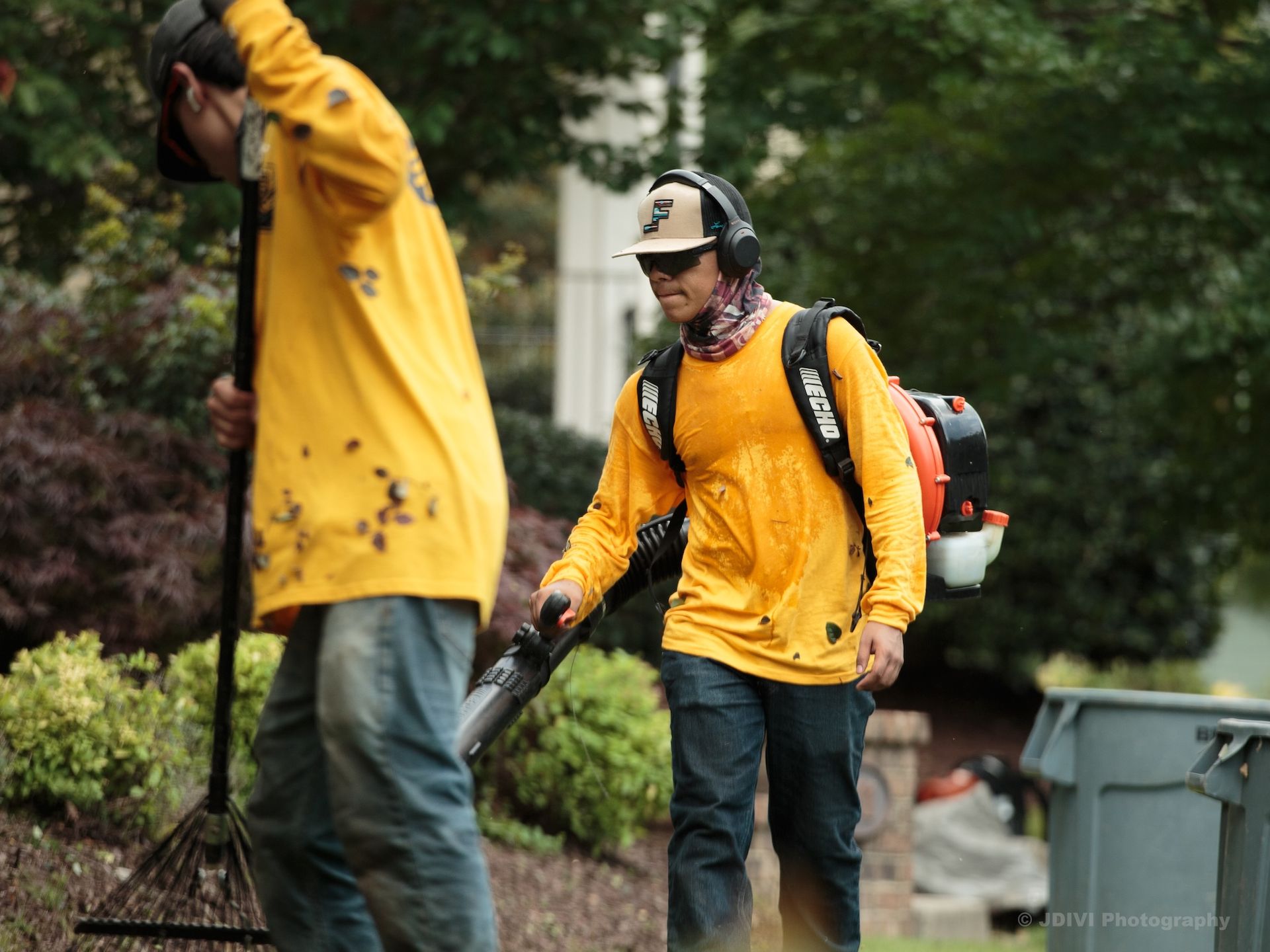 Two landscapers in yellow shirts: one rakes, the other uses a leaf blower on a lawn.