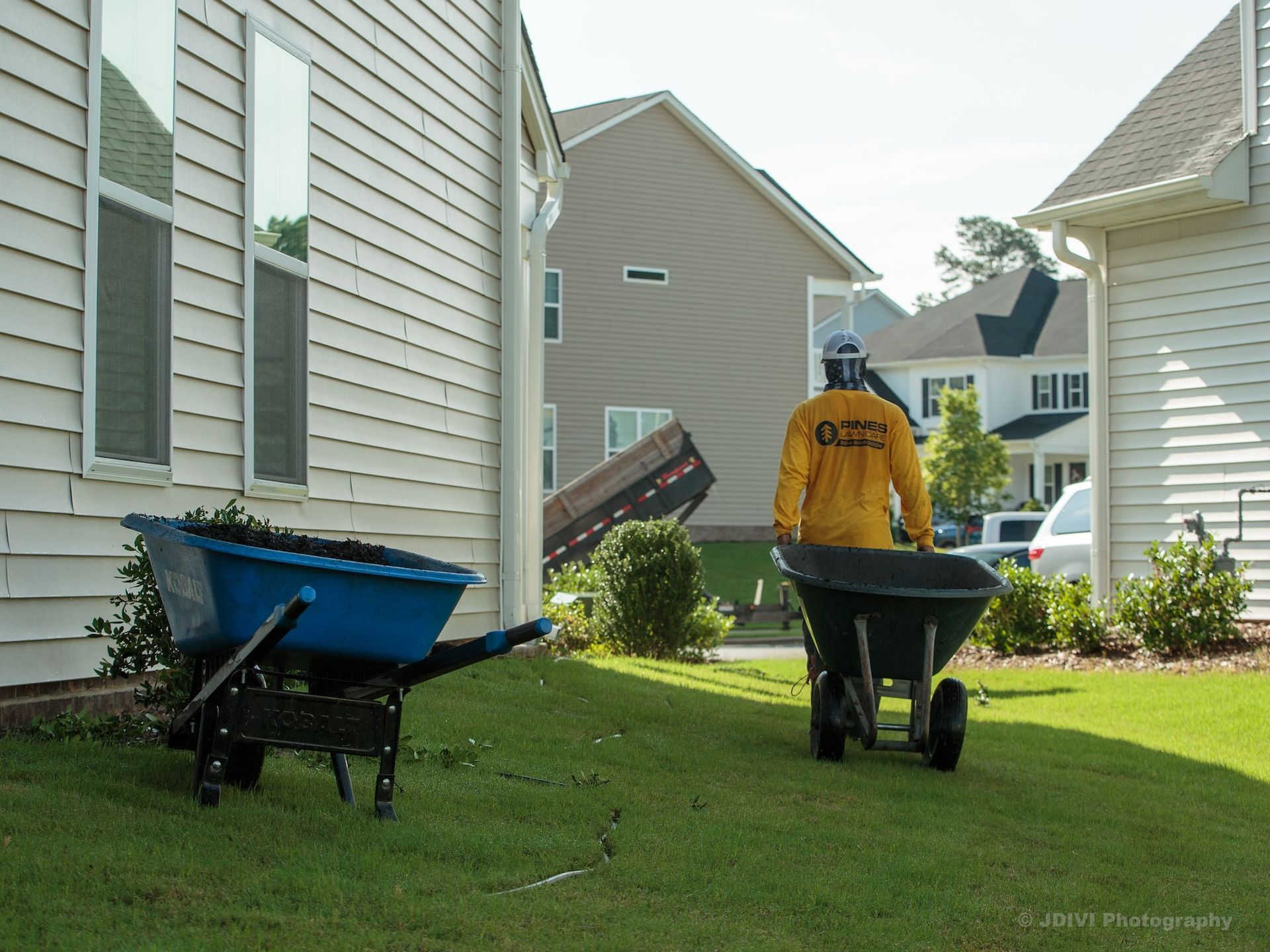 Person in yellow shirt pushing a wheelbarrow on a lawn, another blue wheelbarrow is nearby. Houses in the background.