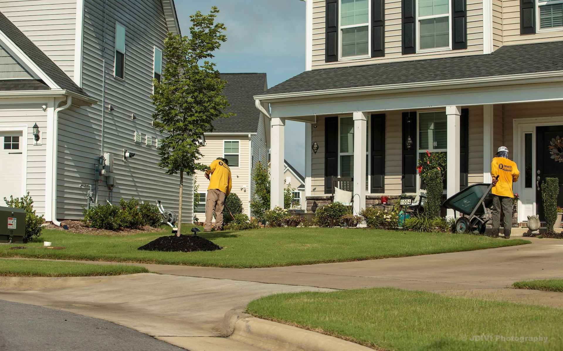 Two landscapers in yellow shirts working on a lawn in front of a house.
