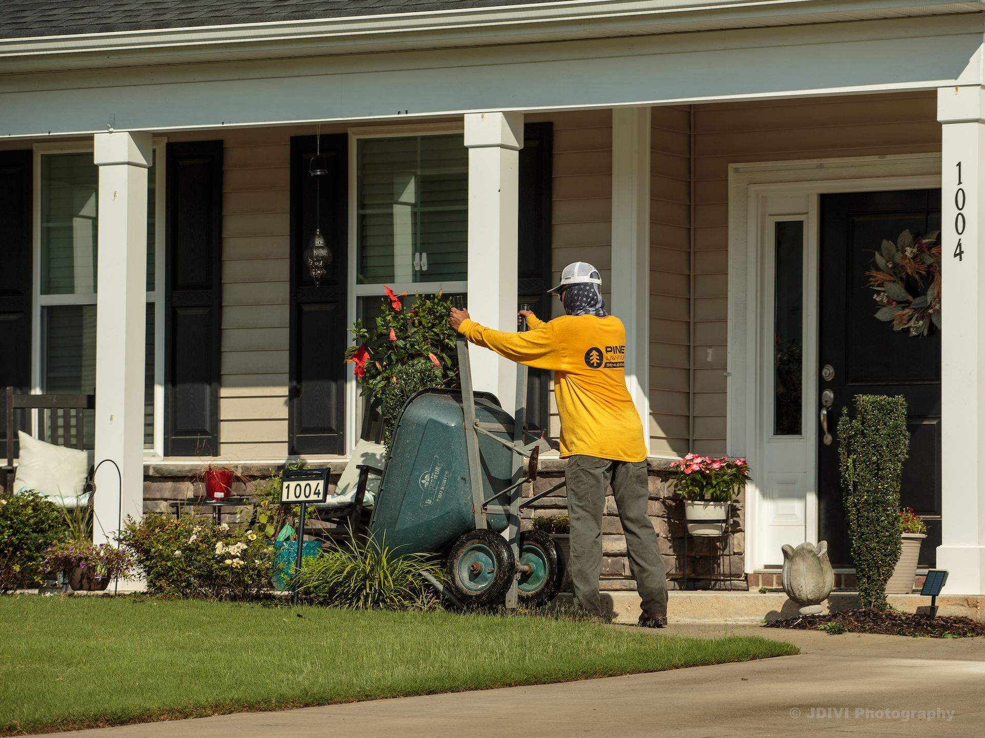 Person in orange shirt operates a lawn edger in front of a beige house with black shutters and door.