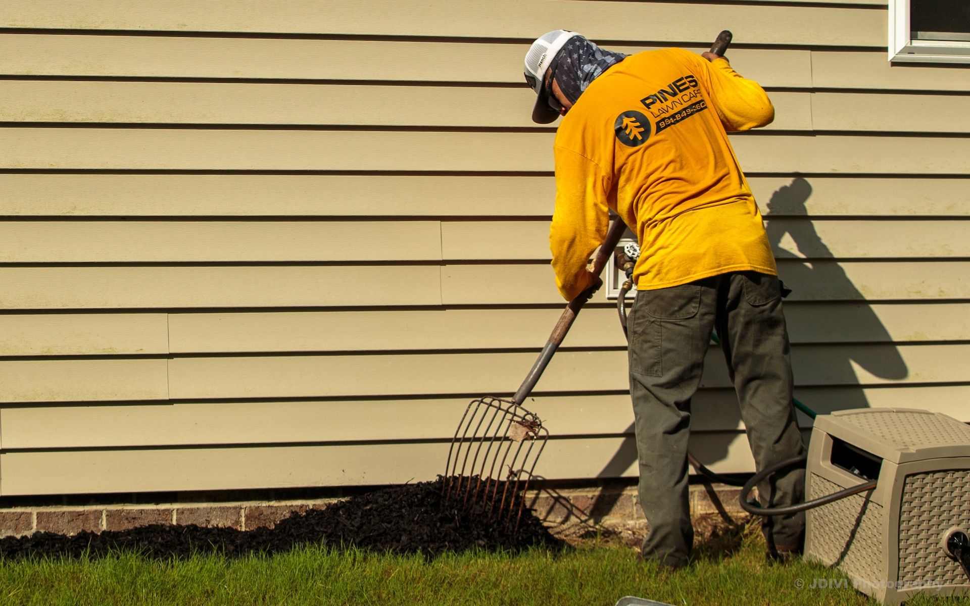 Person spreading mulch with a pitchfork next to a house. Yellow shirt, tan siding, grass.
