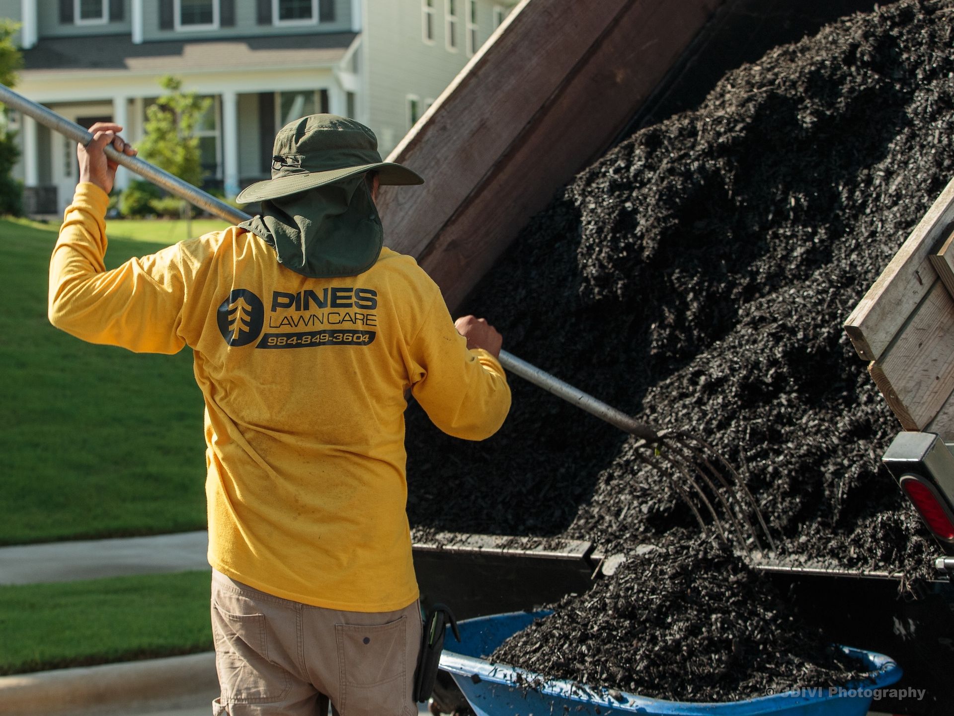 Man in yellow shirt shoveling mulch from truck into a wheelbarrow on a lawn.