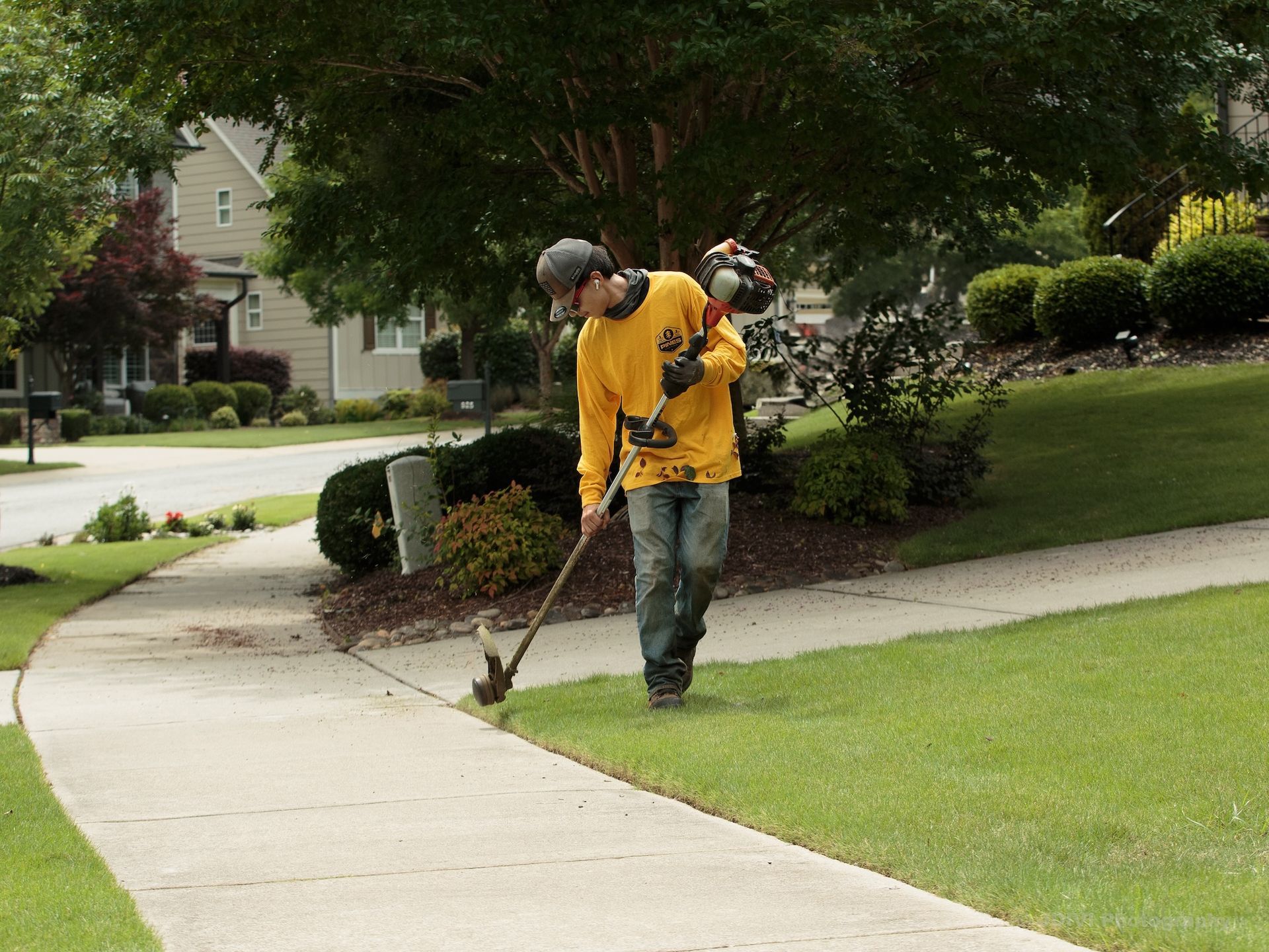 Man trimming grass along a sidewalk with a weed whacker. He wears a yellow shirt, hat, and blue jeans.
