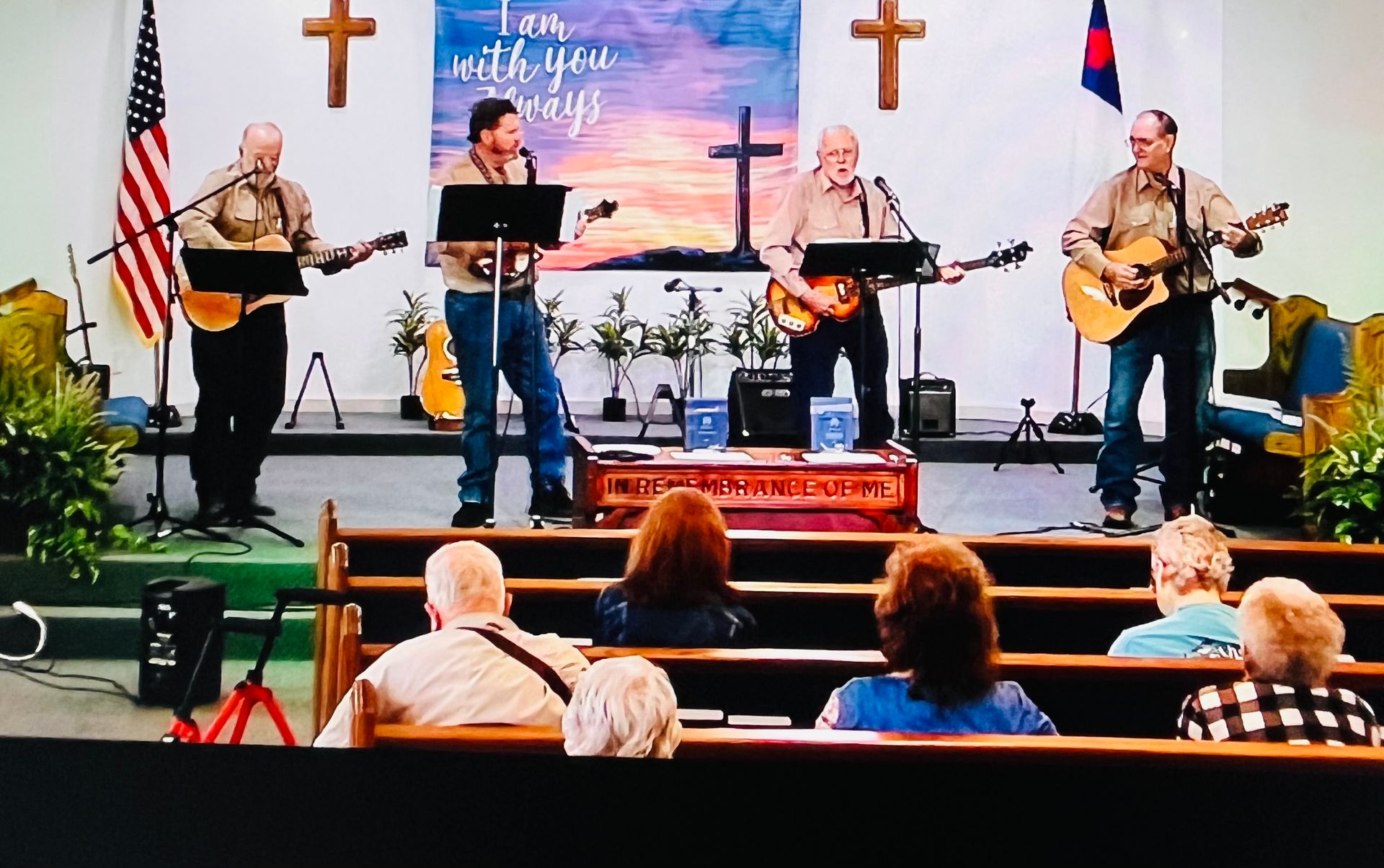 A four-person band performs music on a church stage in front of an audience, accompanied by guitars and microphones.