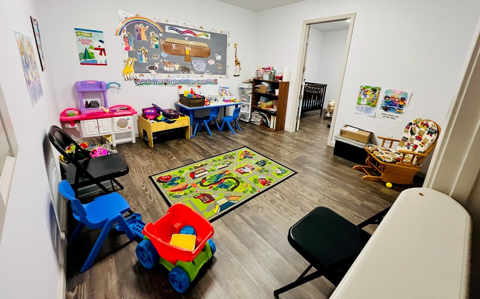 A colorful playroom featuring a rug, toy kitchen, small tables, chairs, a wagon, and a rocking chair on a wood floor.