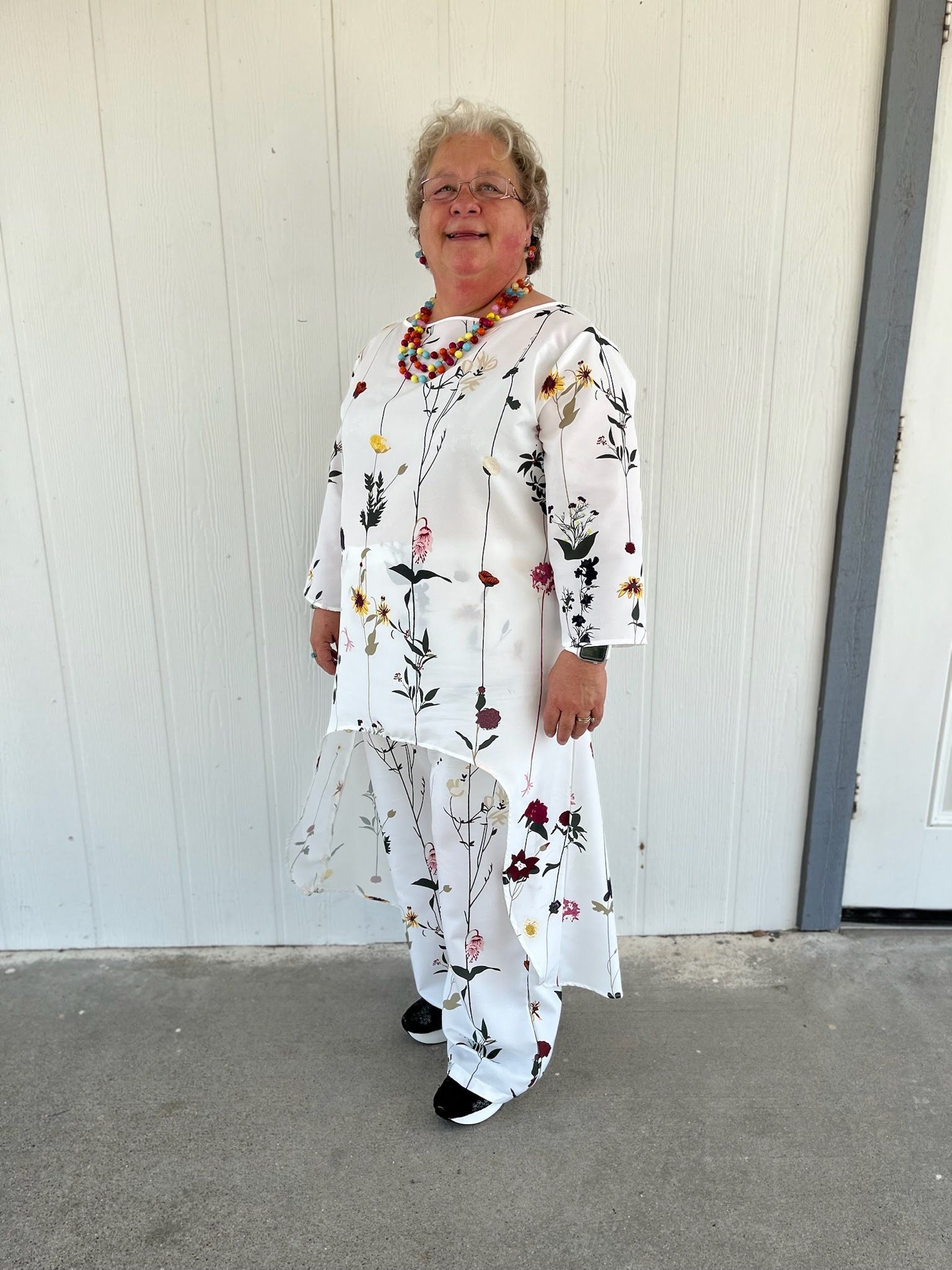 A person with short curly hair smiles while standing outside wearing a white high-low tunic and pants with a floral print.