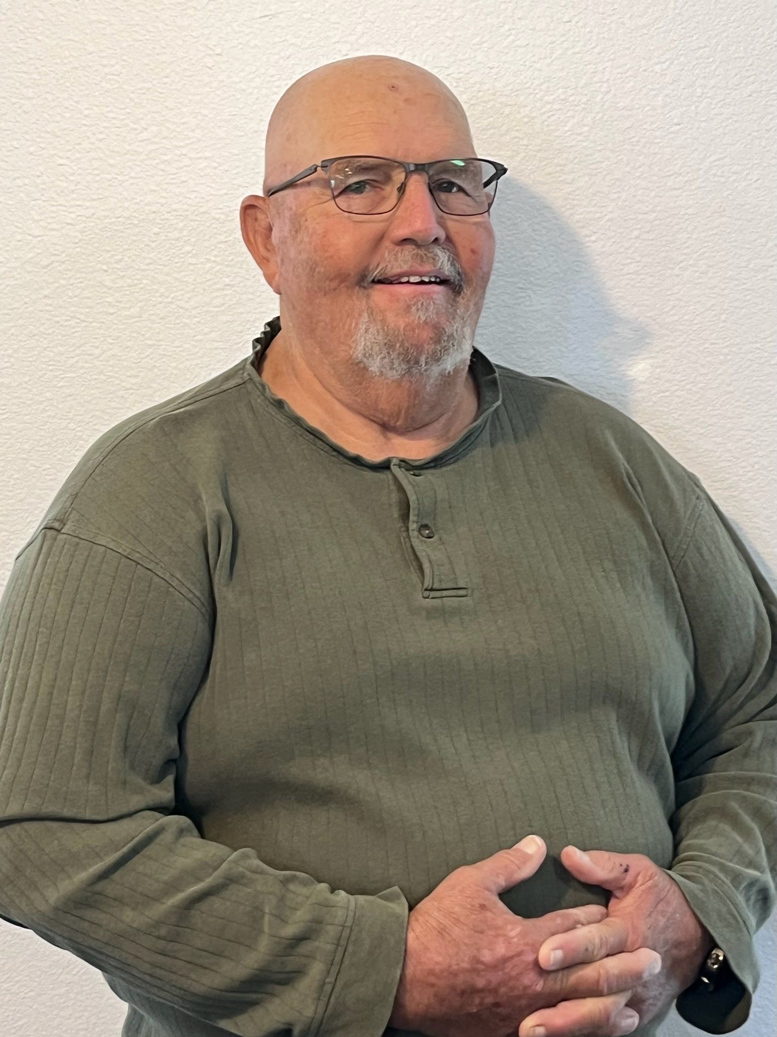 A person with a shaved head and glasses wears a long-sleeved olive shirt, smiling with their hands clasped.