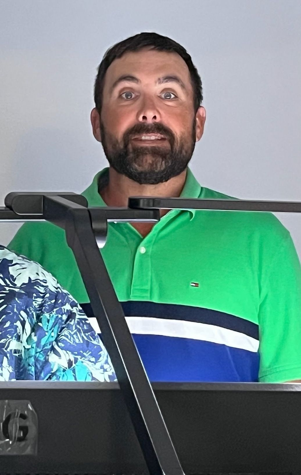 A bearded man in a green, white, and navy blue polo shirt stands behind a black keyboard against a plain background.