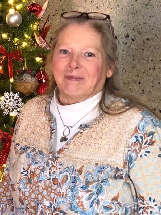 A person with long hair and glasses smiles in front of a decorated Christmas tree with red and gold ornaments.