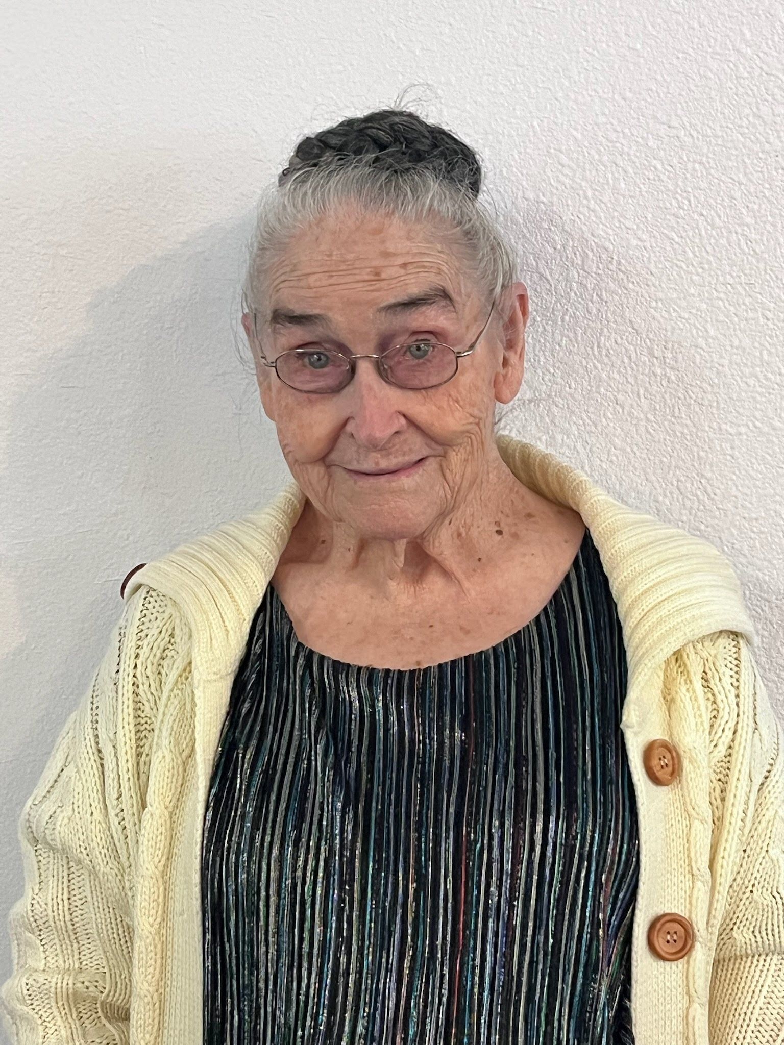 An older person with gray hair styled in a bun, wearing glasses, a striped dark top, and a cream-colored knit cardigan.