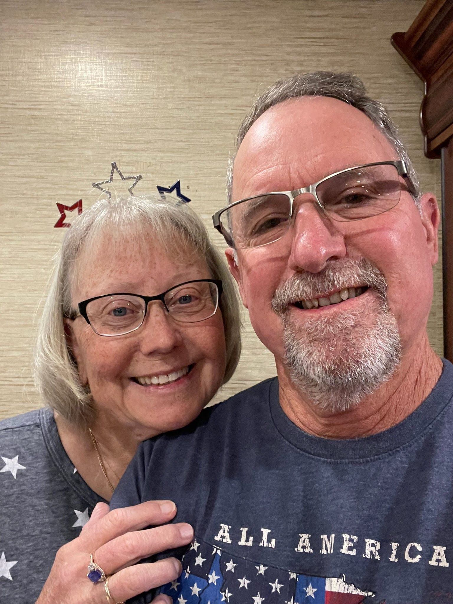 A smiling couple in matching patriotic-themed shirts poses for a selfie against a wall decorated with star cutouts.