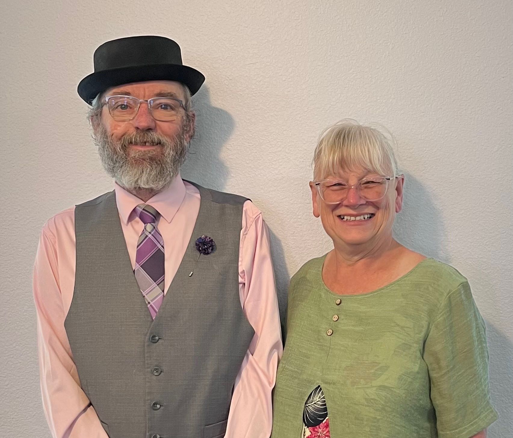 A person wearing a grey vest, tie, and black hat stands next to a smiling person wearing a green top against a plain wall.