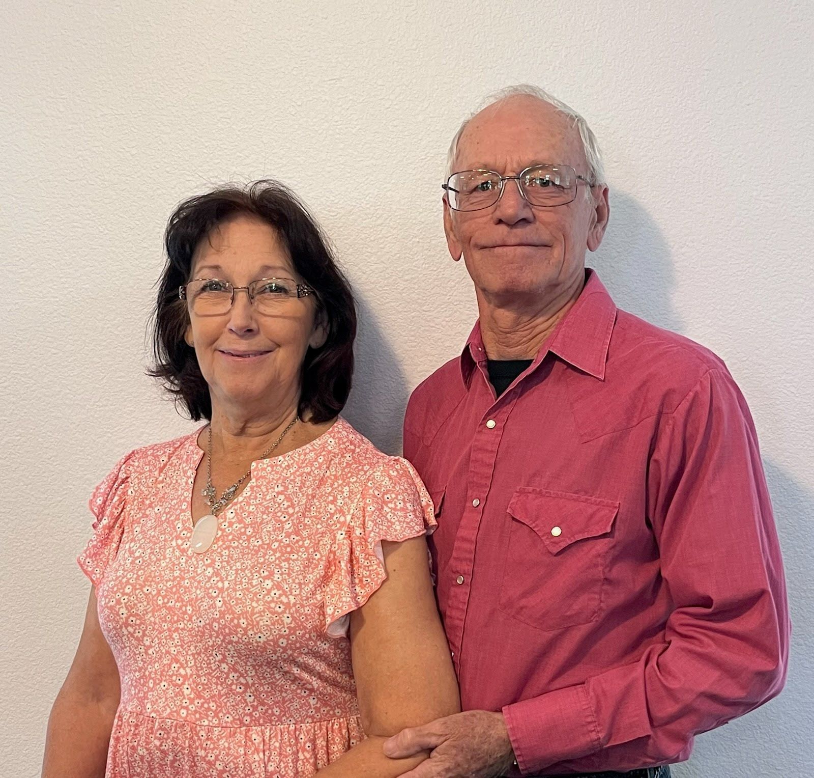 A smiling couple stands side-by-side against a plain white wall; the woman wears a pink floral dress and the man a red shirt.
