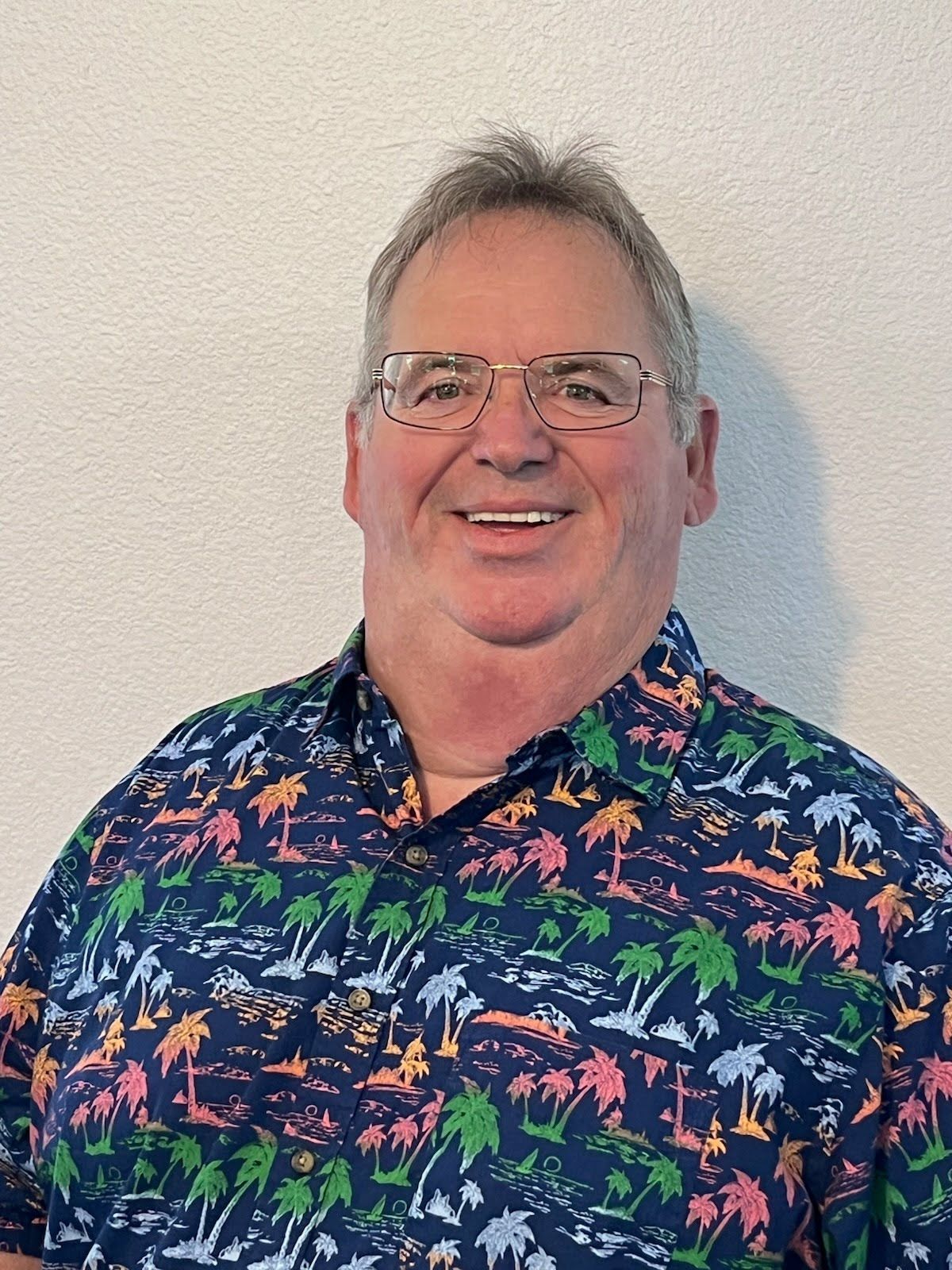 A person with glasses wearing a blue, short-sleeved shirt with a colorful palm tree pattern, smiling against a white wall.