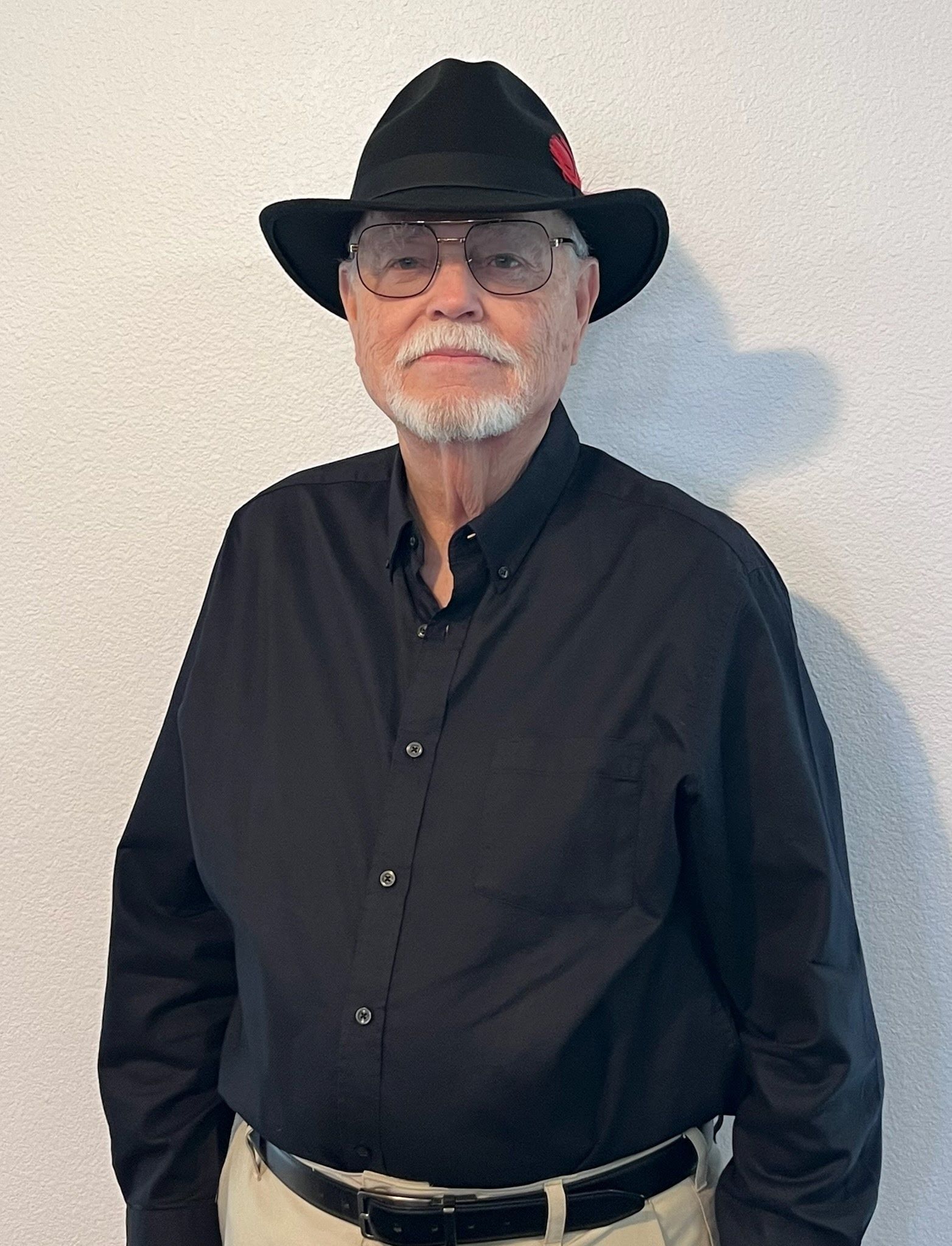 A person wearing glasses, a black fedora with a red accent, and a black button-down shirt stands against a light wall.