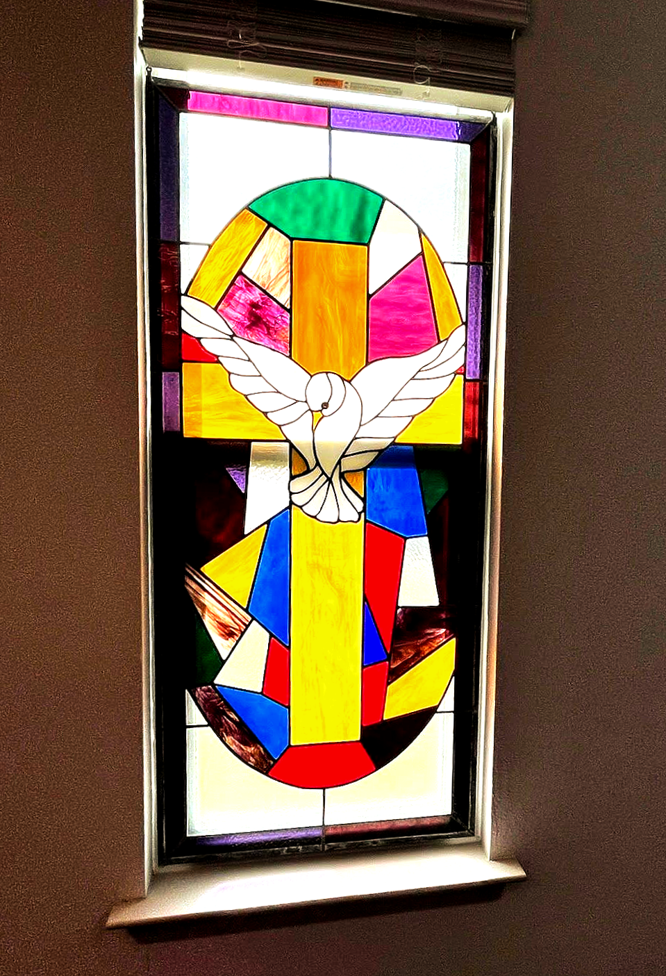 Stained glass window featuring a white dove centered over a yellow cross against a background of multicolored panes.