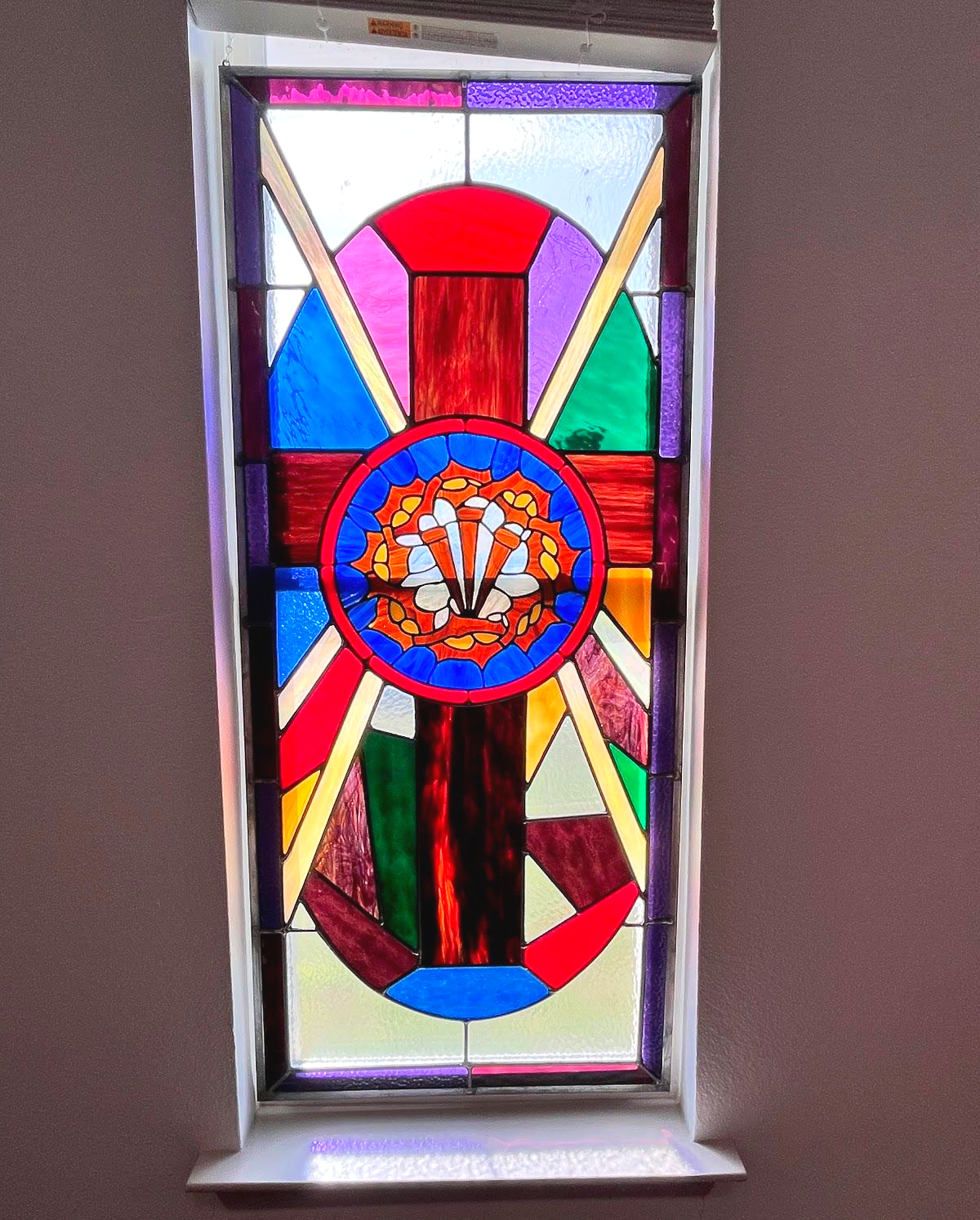 A rectangular stained glass window featuring a central red cross with a circular floral design in multi-colored glass.