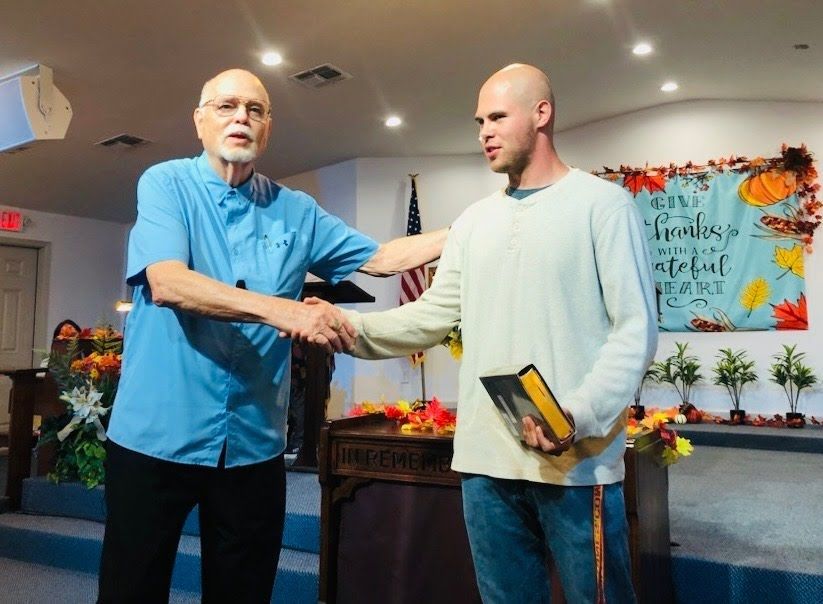 Two men shake hands on a stage in front of a seasonal banner and American flag, one holding a book.