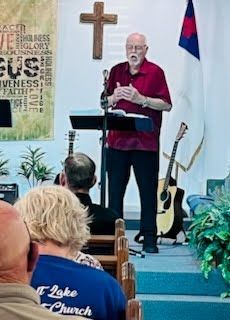 A speaker stands at a podium in a church, addressing an audience with a cross and a Christian flag visible behind them.