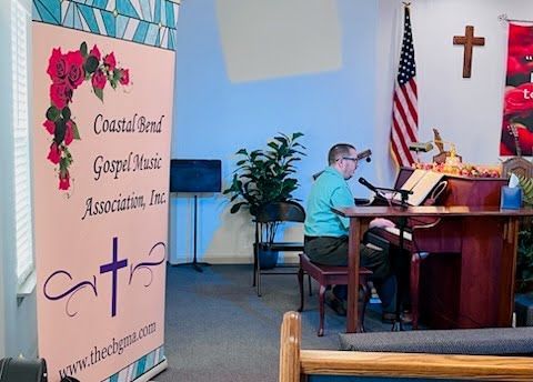 A person plays a piano in a room with a Coastal Bend Gospel Music Association banner, an American flag, and a cross.