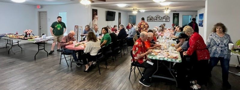 A group of people sitting at tables in a brightly lit indoor space, eating and socializing at a casual event.