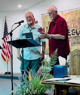Two people stand at a podium in a room with an American flag and a banner featuring religious words, one gesturing.