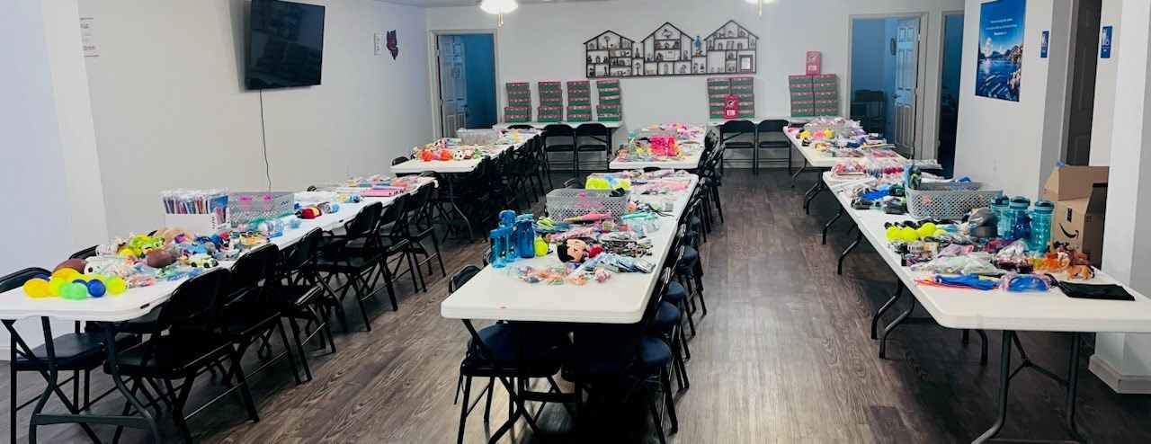Multiple long tables arranged in rows in a room, covered with scattered toys, art supplies, and various small items.