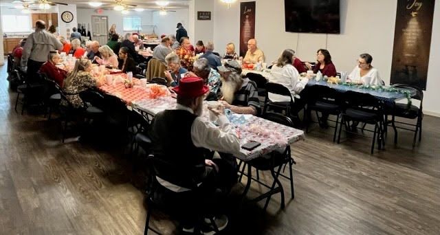 People gather for a meal at long tables decorated with patterned cloths inside a community space or event hall.