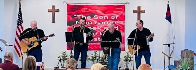 Four musicians stand on a church stage in front of a red banner, playing acoustic guitars and singing into microphones.