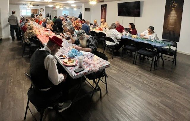 A group of people sit at long tables in a well-lit hall, eating a meal together in an indoor community setting.