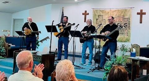 Four men play instruments and sing on a church stage, viewed from the audience with some people applauding.