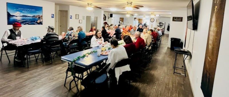 A group of people sits at long tables in a community room, eating and socializing under ceiling fans.
