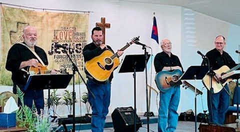 Four musicians in black shirts and jeans perform on stage with guitars, standing before music stands and a church banner.