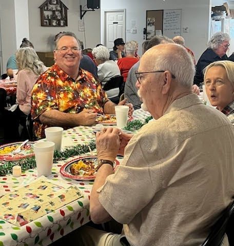 People sit at tables with festive tablecloths, sharing a meal and conversation in a brightly lit community room.