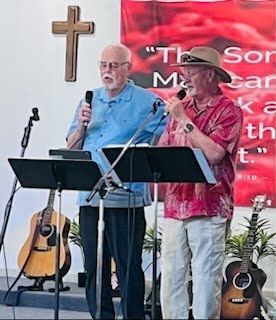 Two people sing into microphones behind music stands in front of a wooden cross and a banner, with guitars on either side.