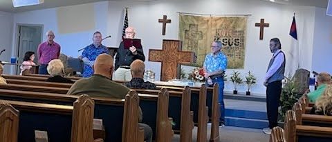 A group of people standing on a stage in a church sanctuary, with a large wooden cross as a backdrop.