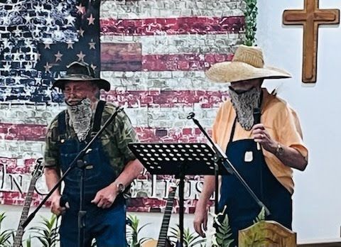 Two performers in overalls and long false beards stand on a stage in front of a US flag backdrop and a wooden cross.