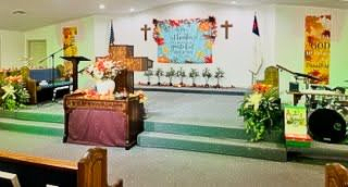The interior of a church featuring a raised stage, a wooden pulpit with floral arrangements, and wall-mounted crosses.
