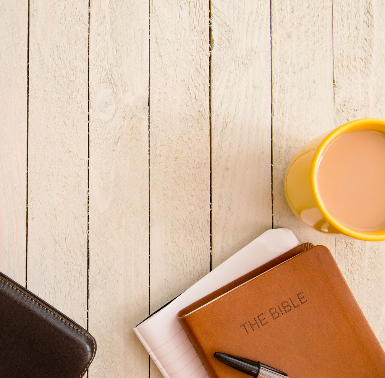 A leather-bound Bible, a notebook, a black pen, and a mug of tea sit on a light-colored wooden table.