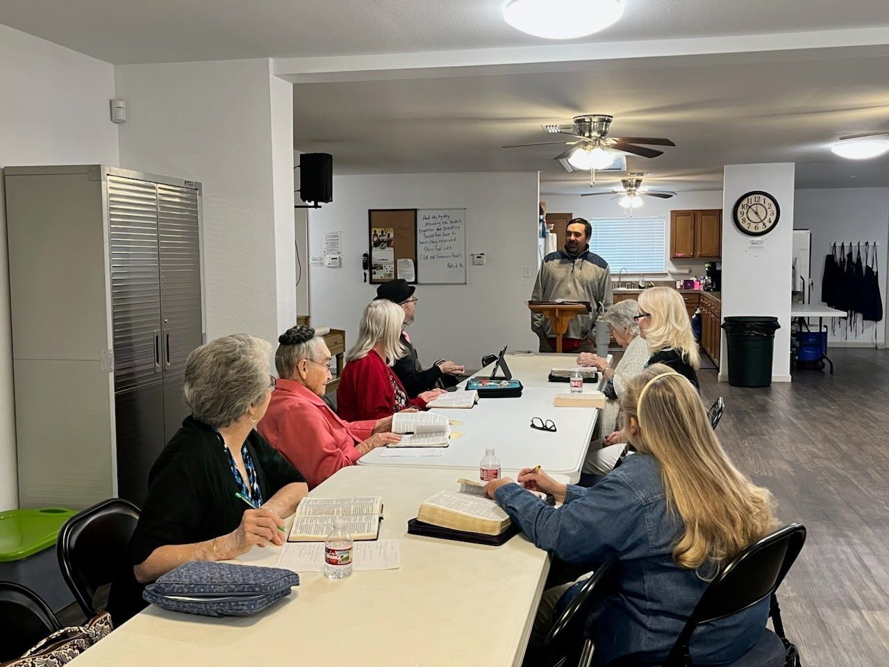 A group of people seated around a long table listening to a man standing at a podium in a community room.