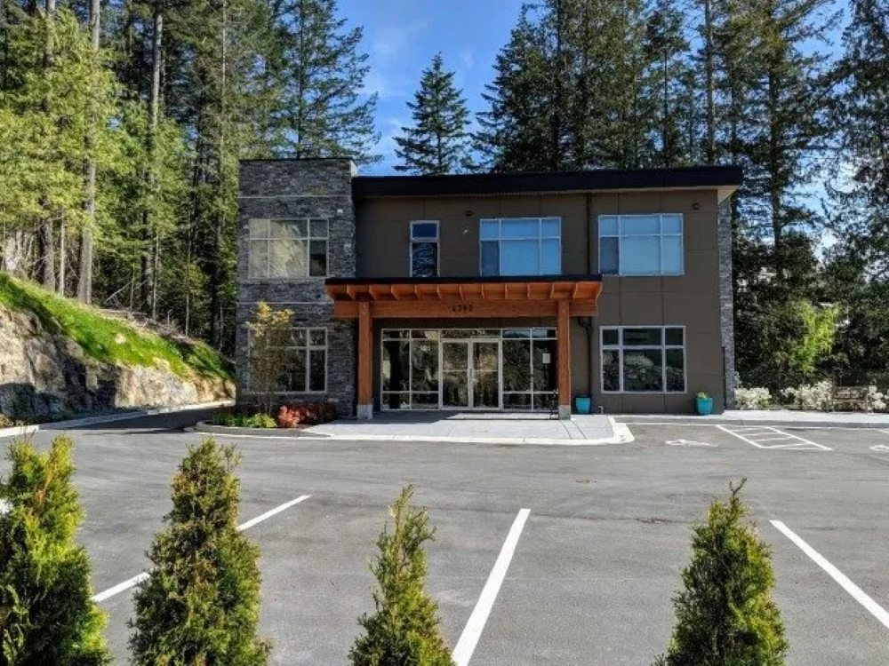 Modern building with stone accents and a wooden entry, surrounded by trees and a parking lot.