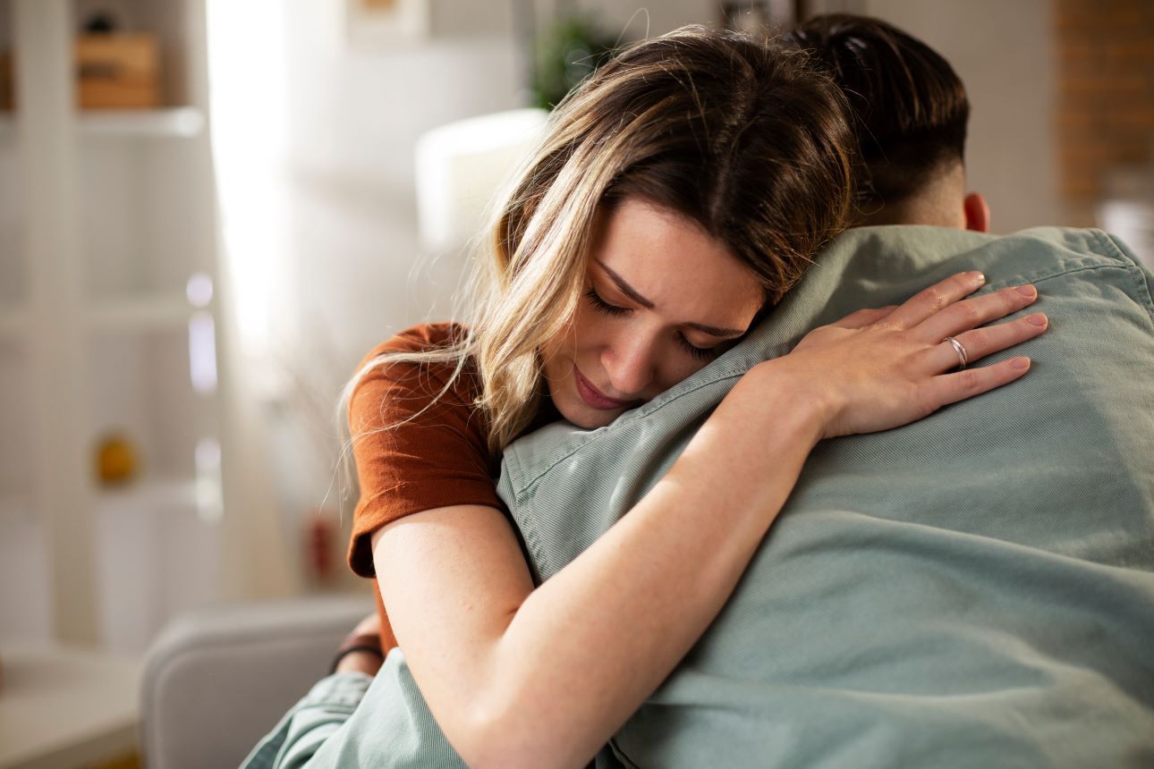 Woman in a brown shirt hugs a person wearing a blue shirt; she appears sad, with eyes closed. Indoors.