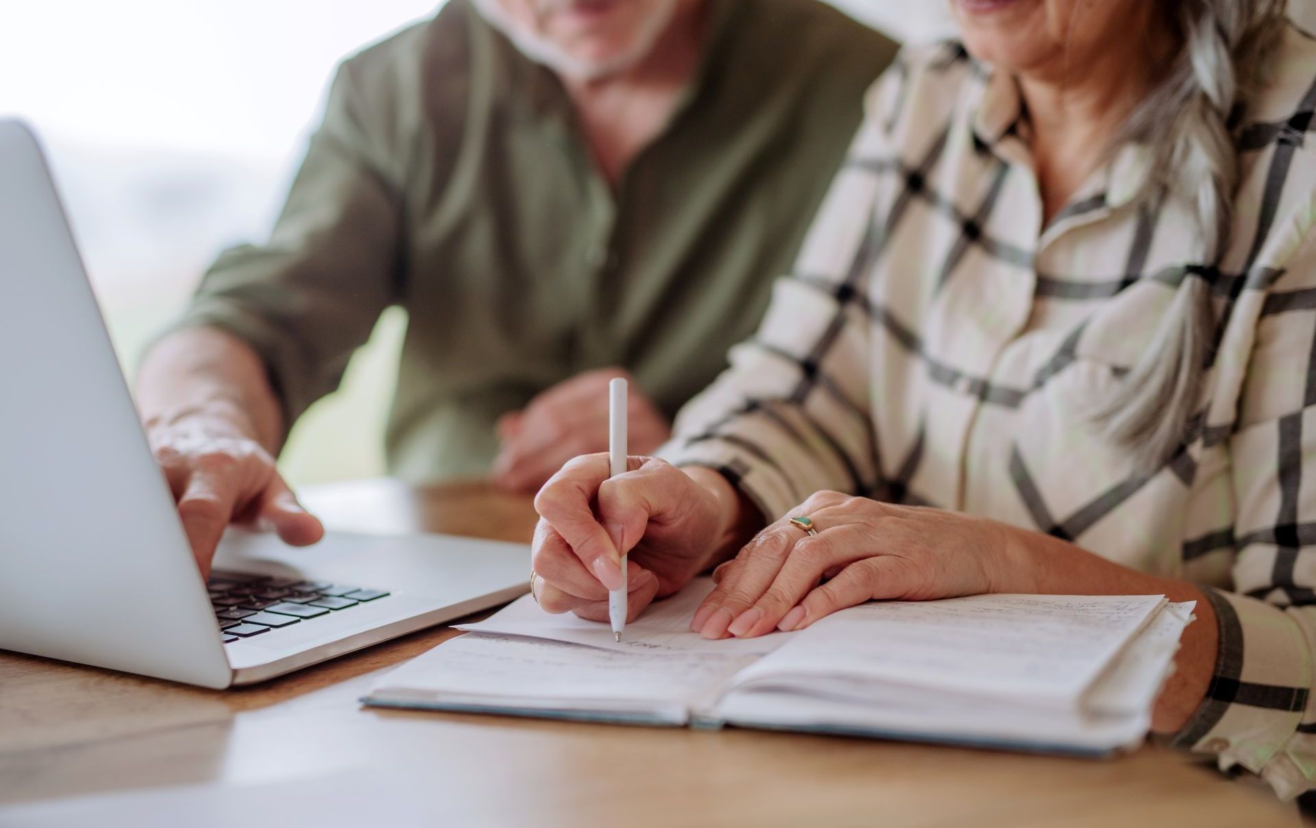 Elderly couple working together, using a laptop and writing in a notebook.