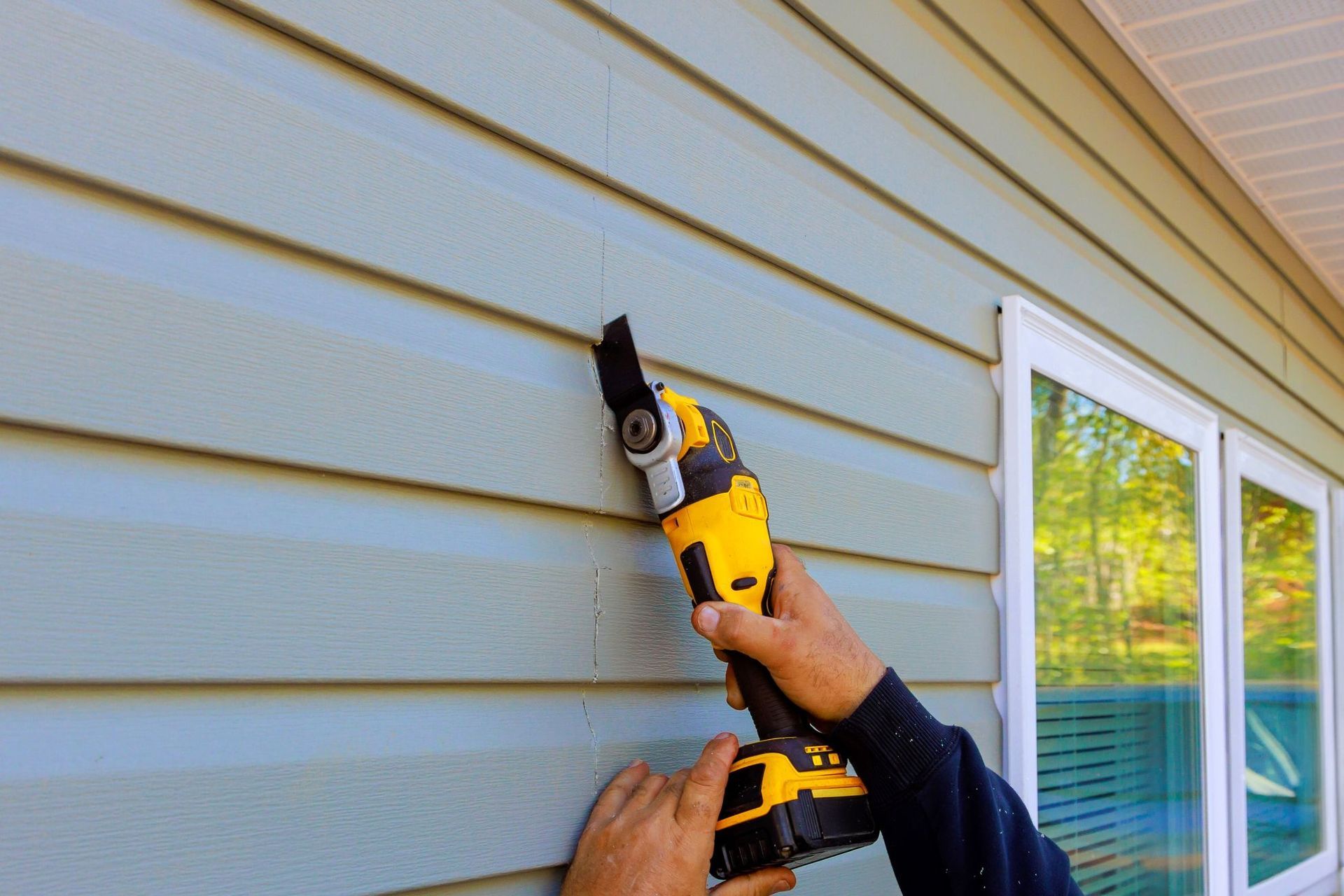 Person using a yellow and black oscillating tool to cut blue siding on a house exterior.