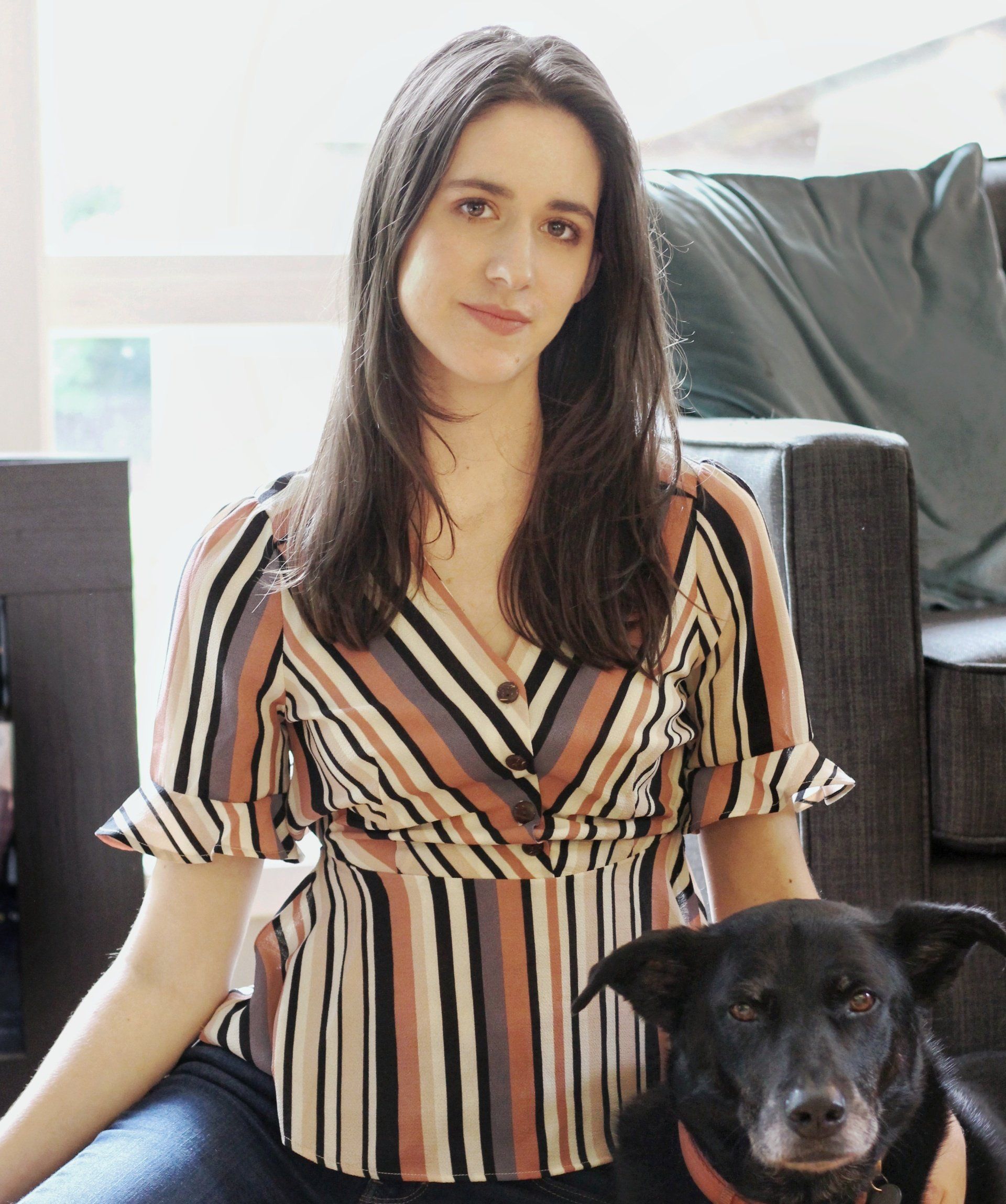 Woman with long dark hair, striped shirt, sitting with a black dog.