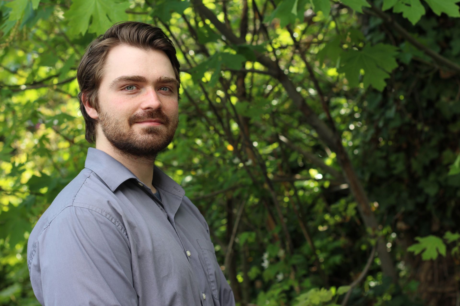 Man with beard, smiles slightly, gray shirt, stands in front of leafy green plants.